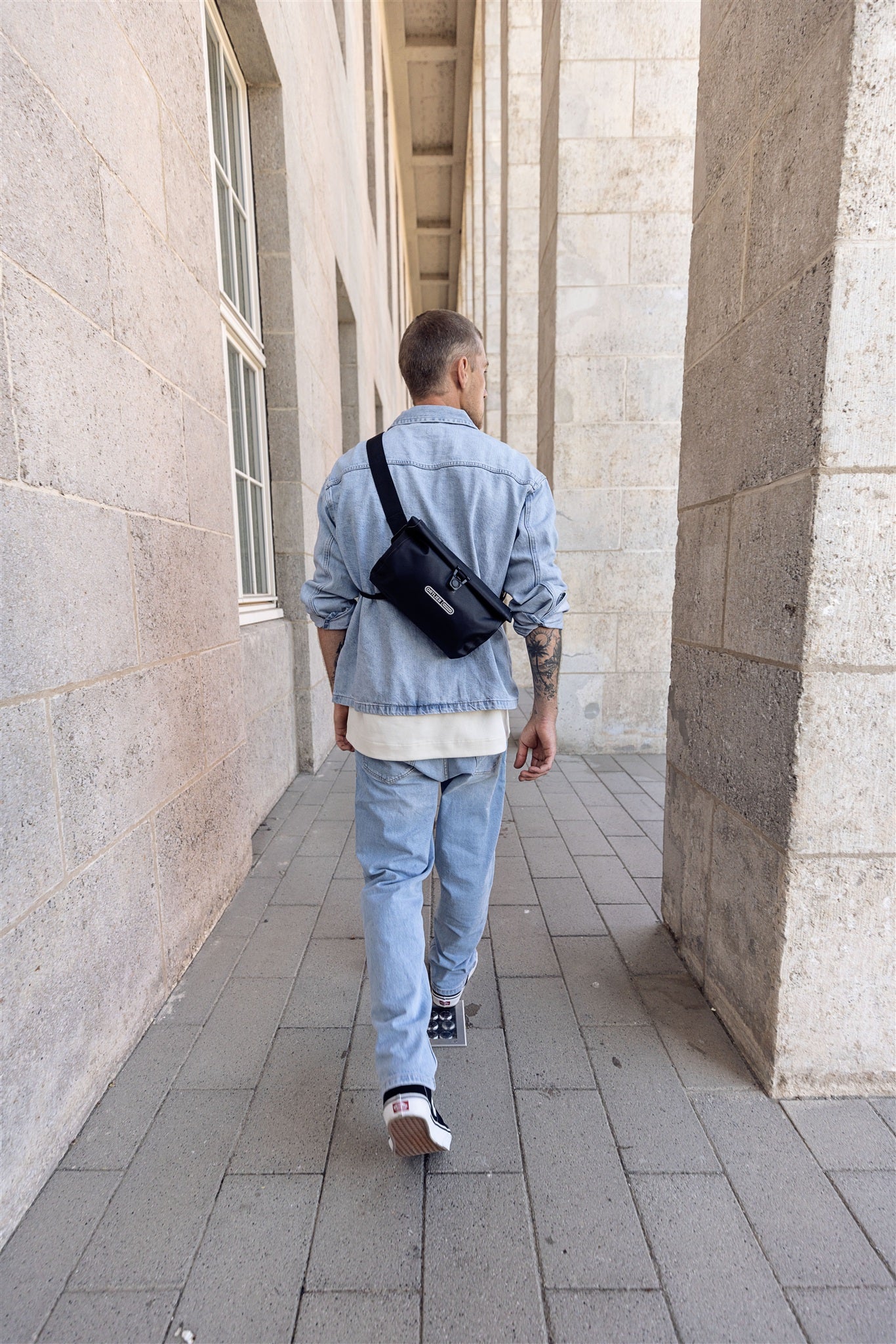 Vista posteriore uomo con tracolla nera ORTLIEB Velo-Sling Flex con chiusura a rotolo mentre cammina tra colonne.